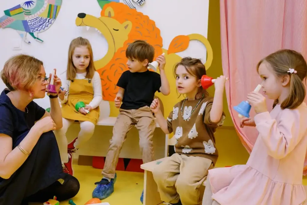 A group of four young children is seated on a bench or stools, holding handbells of different colors and engaging in a playful lesson led by a seated female teacher. The atmosphere is warm and attentive.