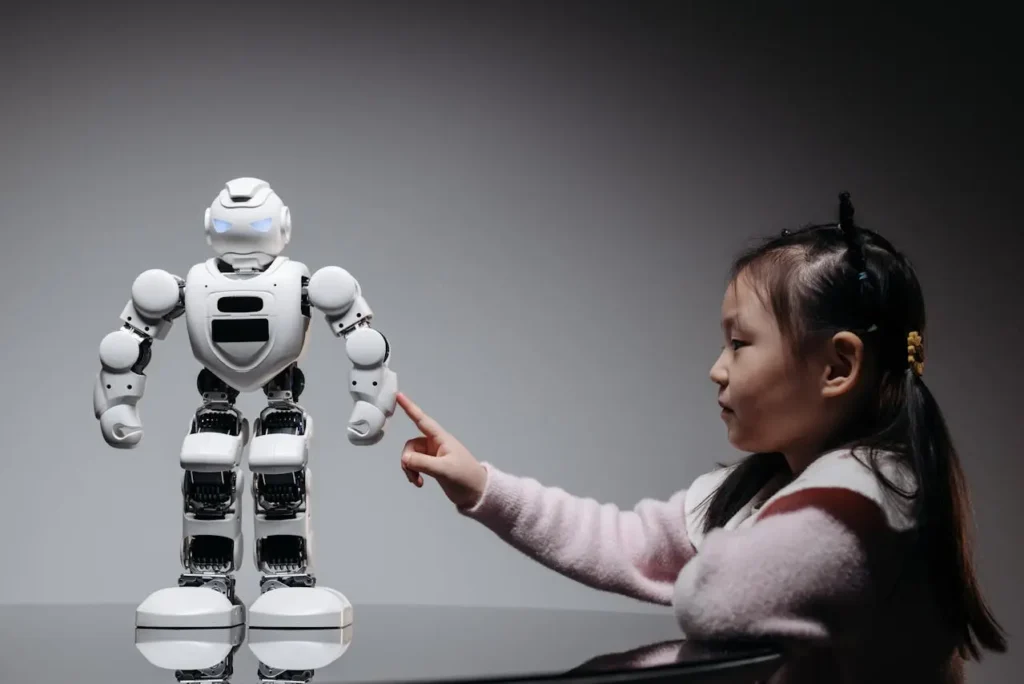 A young Asian girl in a pink sweater is leaning on a dark surface, pointing one finger toward the chest of a large, white humanoid robot with glowing blue eyes in a dark, stylized setting.