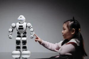 A young Asian girl in a pink sweater is leaning on a dark surface, pointing one finger toward the chest of a large, white humanoid robot with glowing blue eyes in a dark, stylized setting.