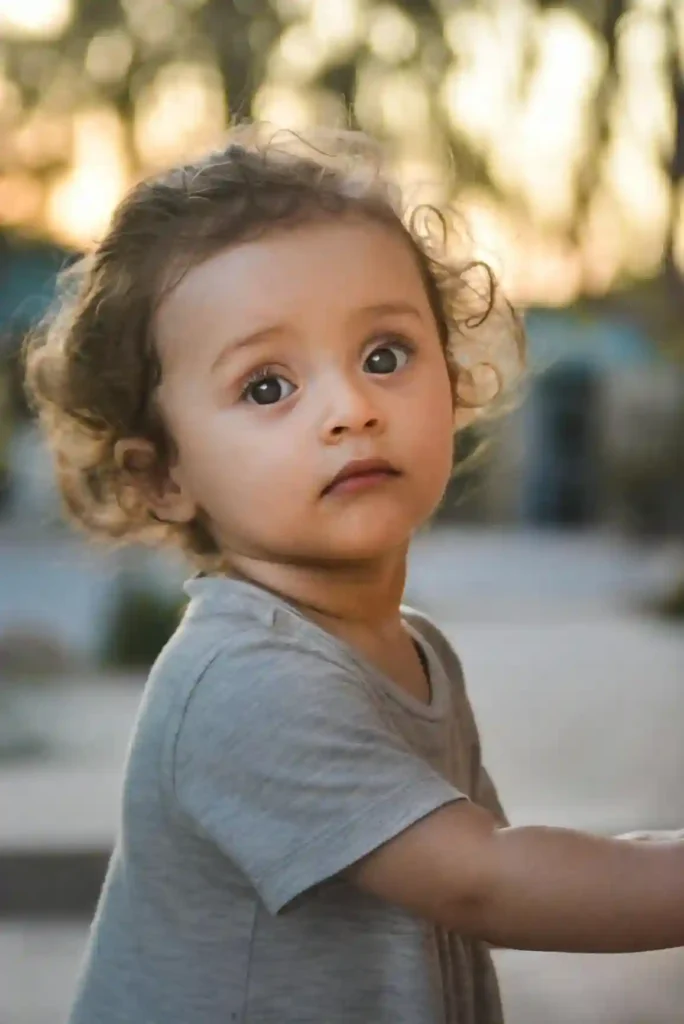 A close-up outdoor portrait of a young toddler with curly brown hair and wide, serious eyes, wearing a simple gray t-shirt. The child's intense gaze suggests a moment of contemplation or focus often encountered when setting boundaries with toddler.