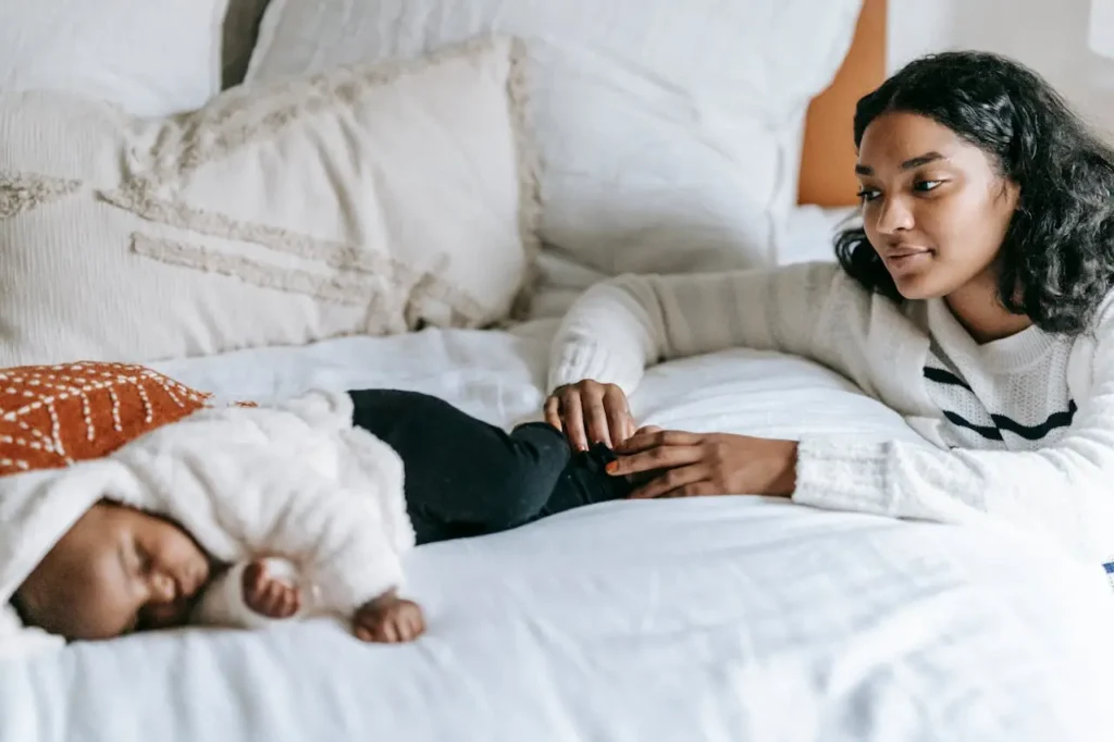 A mother with long dark, curly hair, wearing a white sweater, is lying on a white bed looking affectionately and a bit tiredly at her baby who is sleeping beside her, dressed in a fluffy white hooded sleeper. The image depicts a mother offering comfort during a restless period.