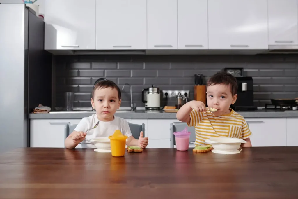Two young twin boys are sitting side-by-side at a dark wooden table in a modern kitchen. They are eating a meal or snack from matching white bowls with spoons. One boy is wearing a yellow striped shirt and the other a white shirt.