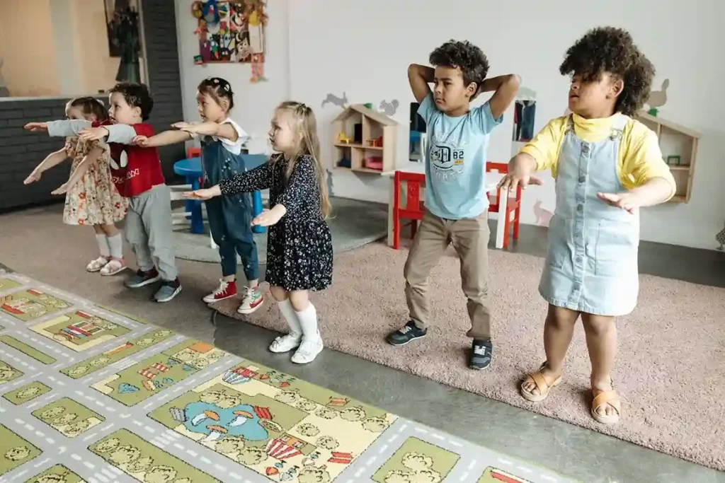 A group of six diverse toddler and preschool-aged children is standing in a line in a brightly lit room, performing a movement, possibly raising their arms out to the sides or up to their heads as part of a cooperative game. This activity is ideal for teaching Social Skills Games for Toddlers and Preschoolers.