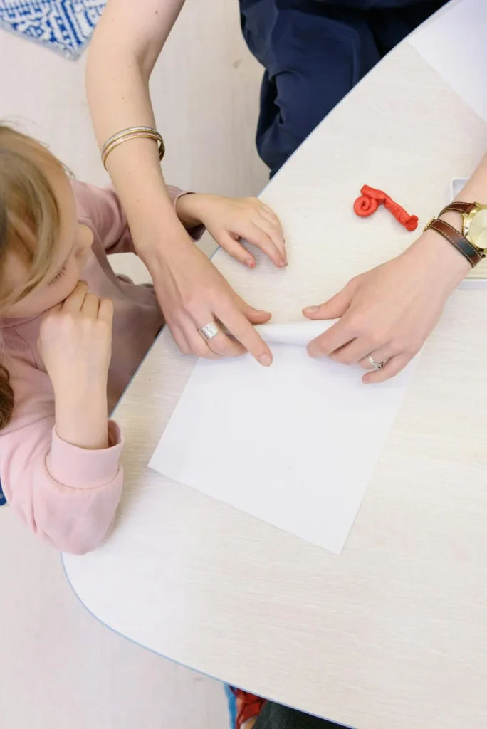 An overhead shot of an adult's hands (wearing a watch and rings) guiding a young girl's hands to fold a piece of white paper on a table. This fine motor activity is often paired with songs rhymes and fingerplays that boost language skills.