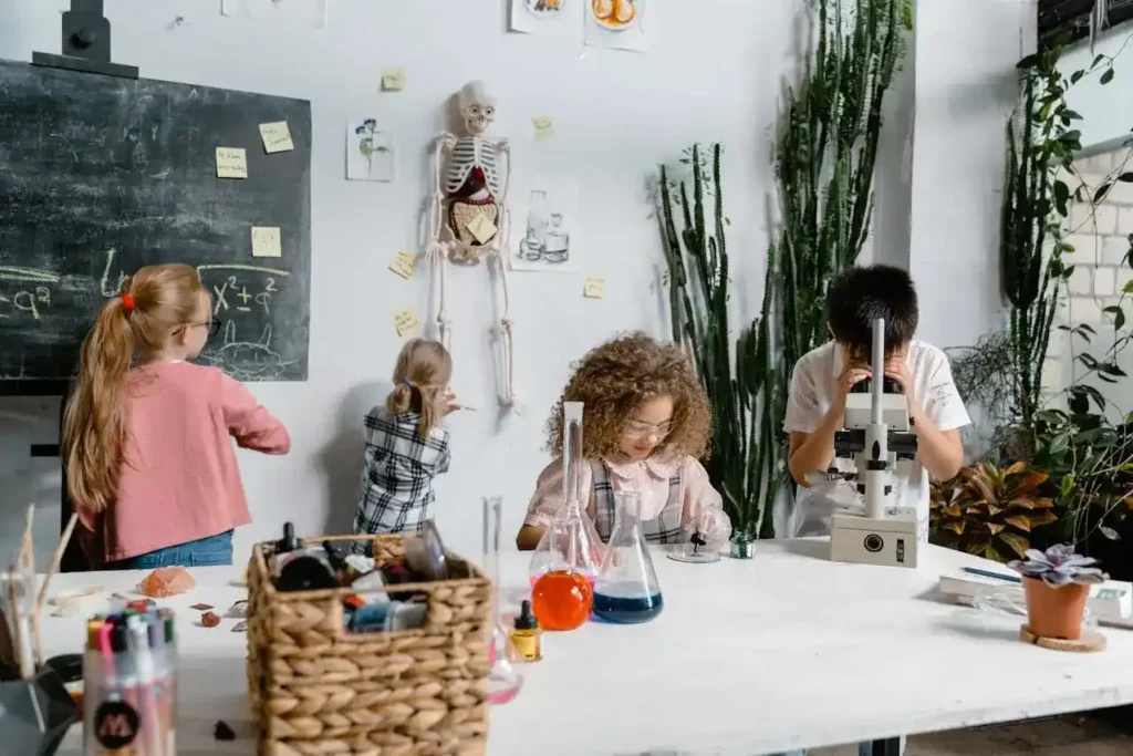 Four children in a science-themed classroom: a boy is looking through a microscope, a girl is using beakers and liquids, and two other children are writing on a blackboard and examining a model skeleton.
