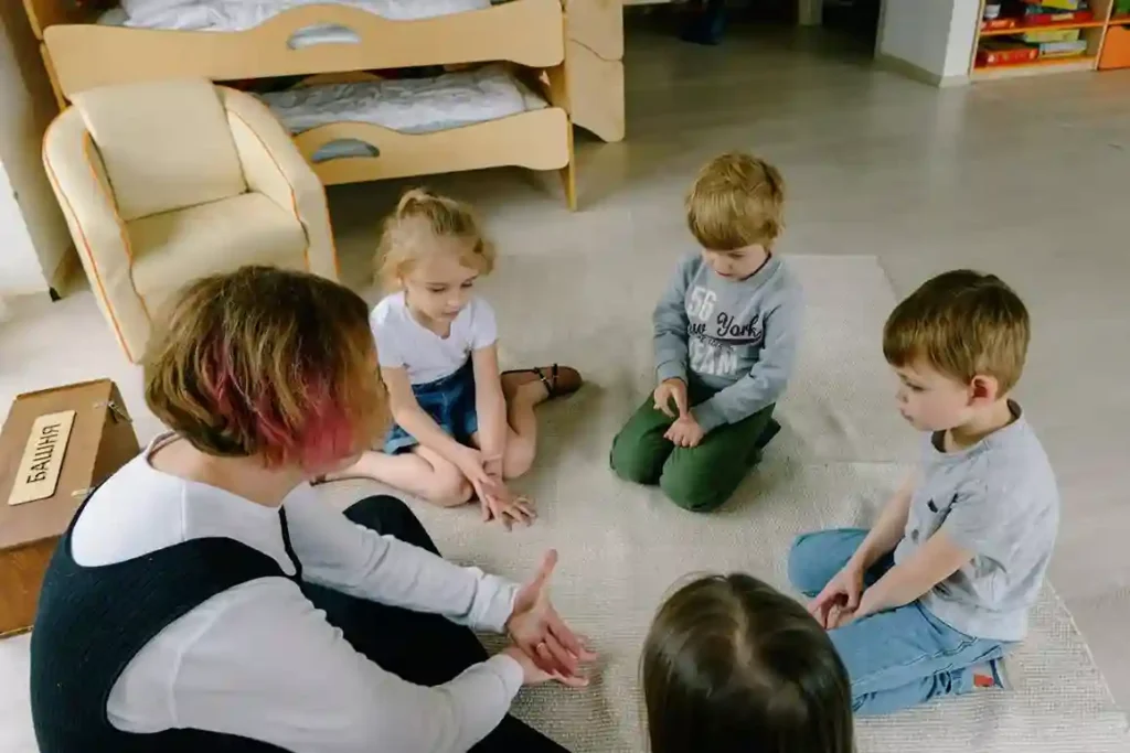 A teacher with pink-tipped hair is seated on the floor with four young children in a circle, engaged in a conversation or story time activity in a brightly lit classroom. This group setting helps in supporting early friendships in preschool and daycare.