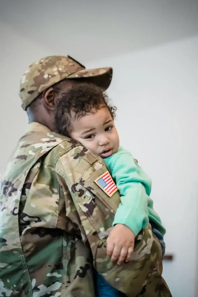 A close-up vertical shot of a person in a U.S. Army uniform and cap holding a young toddler, who is resting her head on the service member's shoulder and looking sadly at the camera. An American flag patch is visible on the uniform.