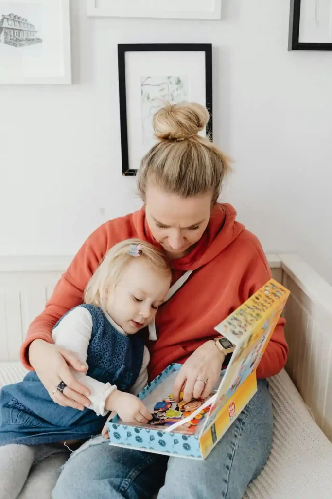 A mother wearing an orange hoodie is sitting and hugging her young daughter, who is wearing a denim dress. They are looking down at a box of puzzle pieces together, an activity ideal for teaching cause and effect for toddlers.