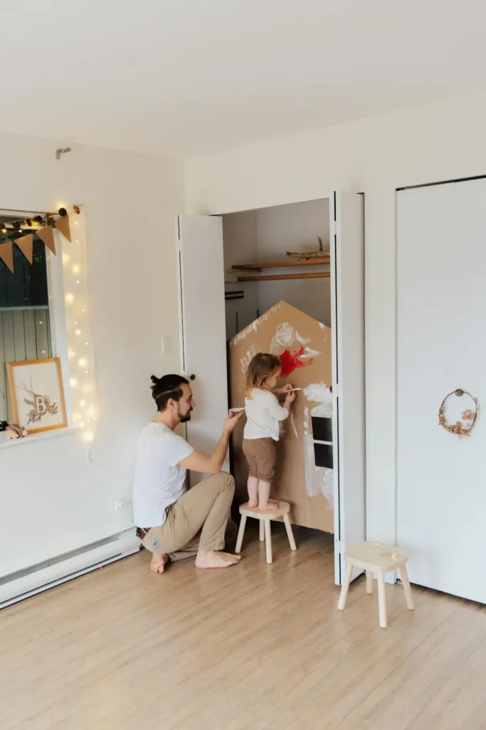 A man with a beard and a topknot is crouching down with his young toddler son, who is standing on a stool, as they both paint the white exterior of a large, cardboard playhouse built inside a closet or room corner. This activity involves Teaching Toddlers to Share and Take Turns with the paintbrush.