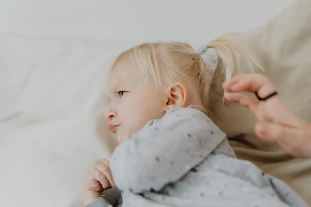 A close-up side profile of a young girl with blonde hair tied in pigtails, lying in bed under a gray patterned blanket. She has a firm, slightly resistant expression and is looking away from a partially visible adult hand trying to comfort her.