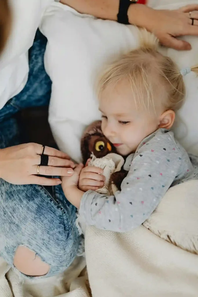 A close-up of a young girl with blonde pigtails lying down and holding a small brown stuffed animal, tucked under a blanket. An adult hand is gently resting near her hand, providing comfort during a quiet time.