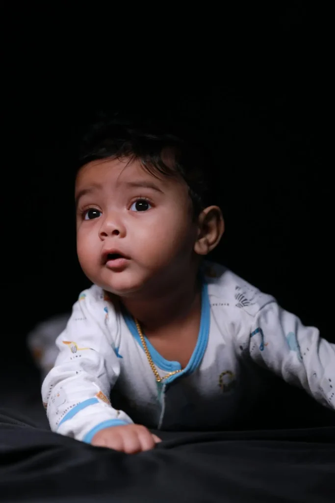 A close-up portrait of a wide-eyed baby, lying on their stomach on a dark surface and looking upward. The lighting is focused only on the baby, emphasizing the dark environment, which is typical of toddler night wakings and early rising.