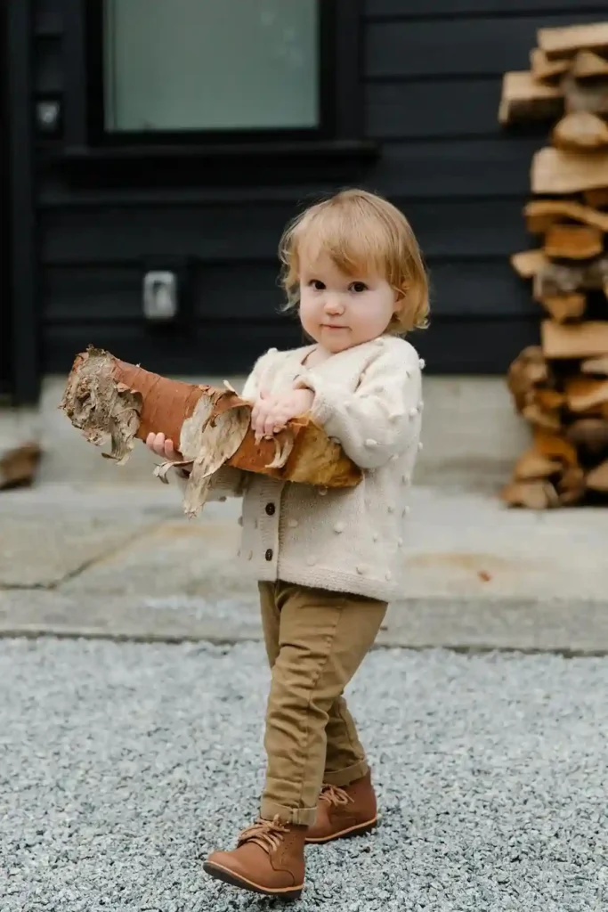 A cute blonde toddler, wearing a cream-colored cardigan with pom-poms and brown pants, is standing outdoors on a gravel path, happily holding a large, textural piece of birch tree bark.