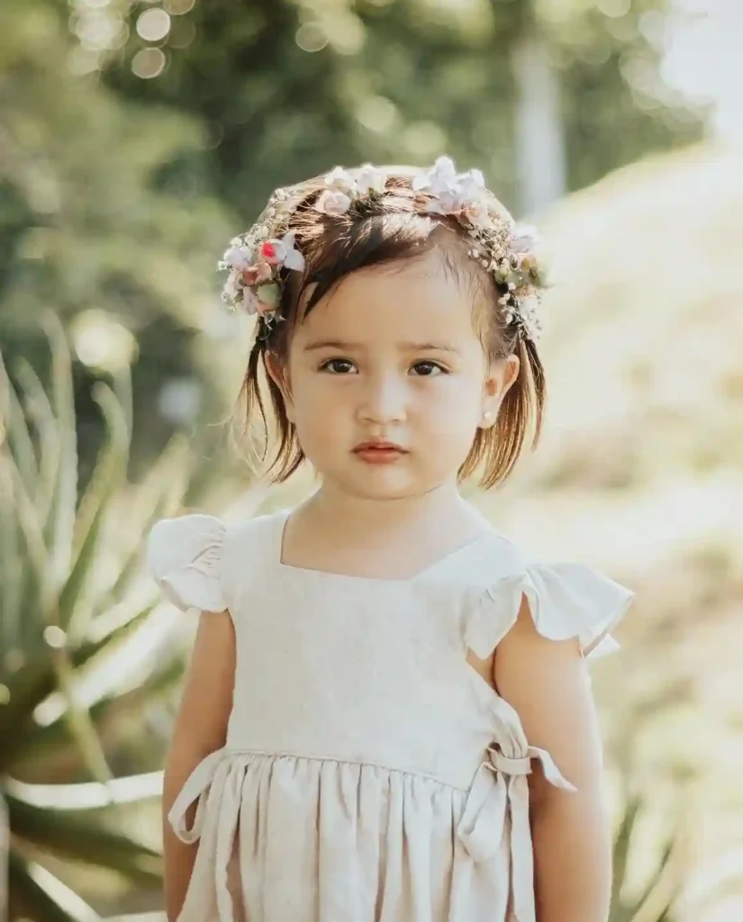 A close-up portrait of a serious-faced toddler girl with short brown hair, wearing a white sleeveless dress and a delicate crown of flowers in her hair, looking directly at the camera with a firm expression.