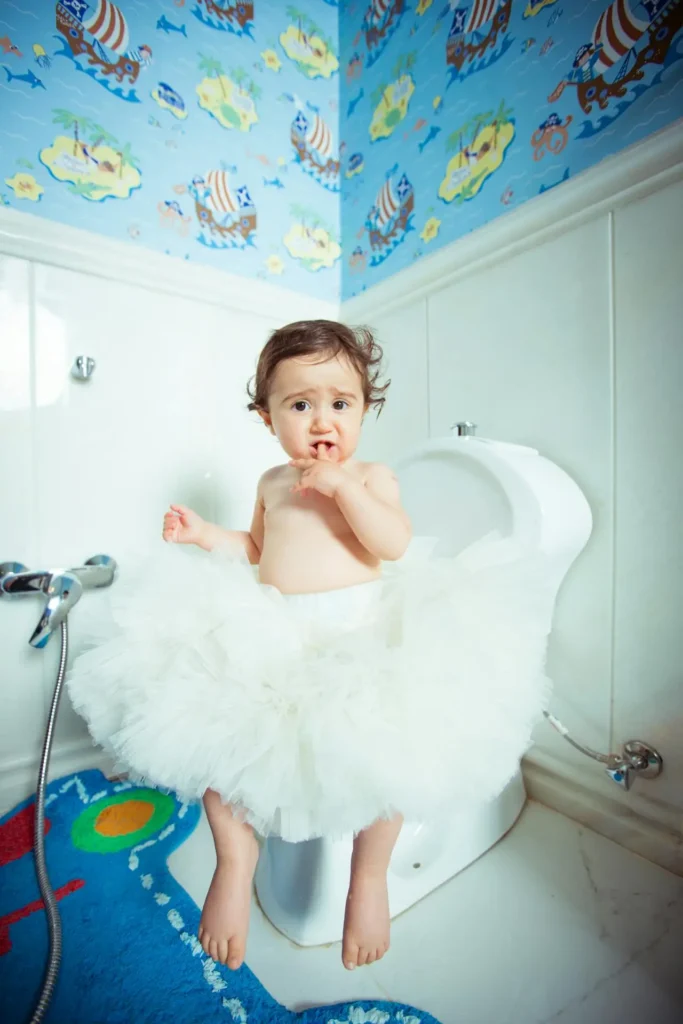 A cute baby or young toddler, wearing only a large white tutu skirt, is sitting on a regular-sized toilet, looking directly at the camera with a surprised expression. The bathroom is decorated with brightly colored wallpaper.