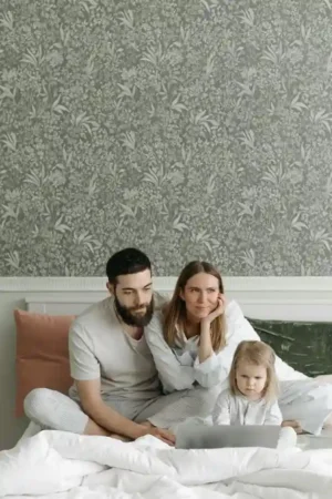 A mother, father, and young toddler are sitting together on a large bed with floral wallpaper behind them, looking down at a laptop screen in front of them. The toddler appears attentive or stressed by the screen content.