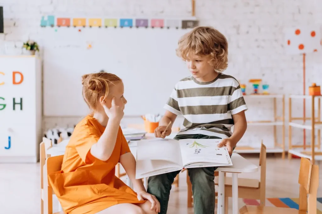 Two young children, a blonde girl in an orange dress and a boy in a striped shirt, are sitting at a small white table in a bright classroom. The boy is holding an open book and speaking to the girl, who is listening intently and touching her ear.